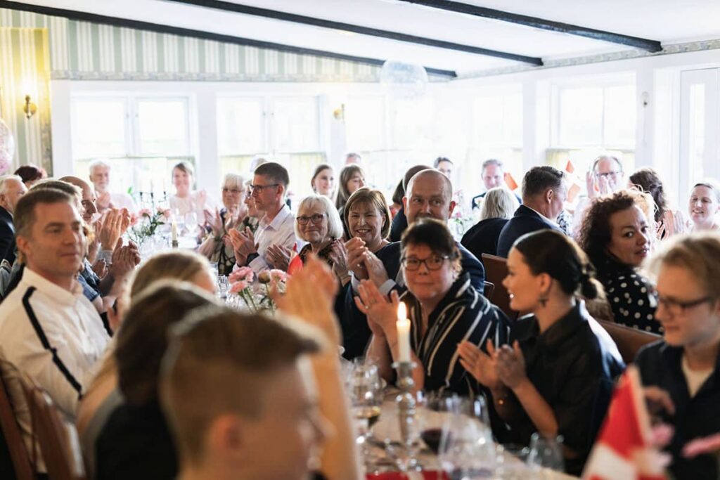 Guests at a confirmation celebration laughing and clapping around the decorated table with Danish flags