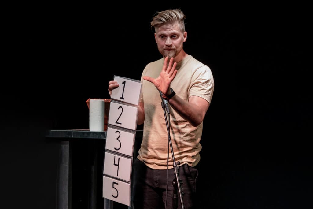 Anders Andresen laver trylleri og stand up på en mørk scene med fem nummererede kort ved mikrofonstativet
